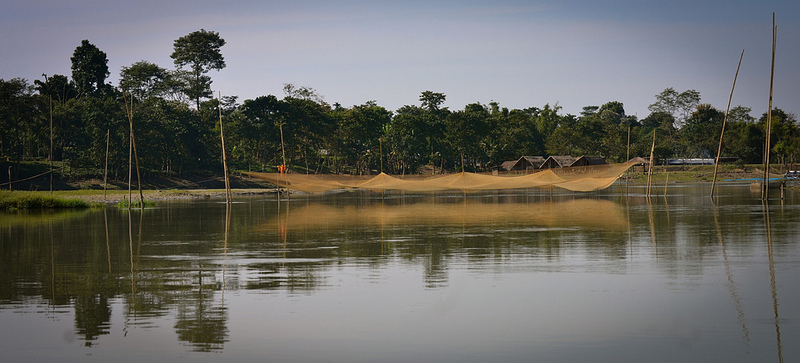 A huge fishing net standing in the middle of the beel with houses and trees in the background 